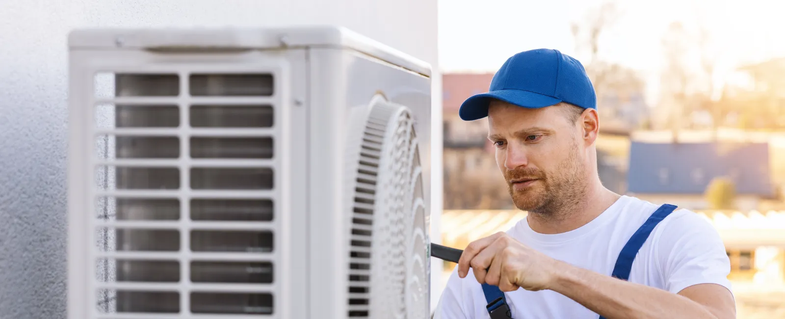 Technician in blue overalls and cap repairing an outdoor air conditioning unit with a screwdriver.