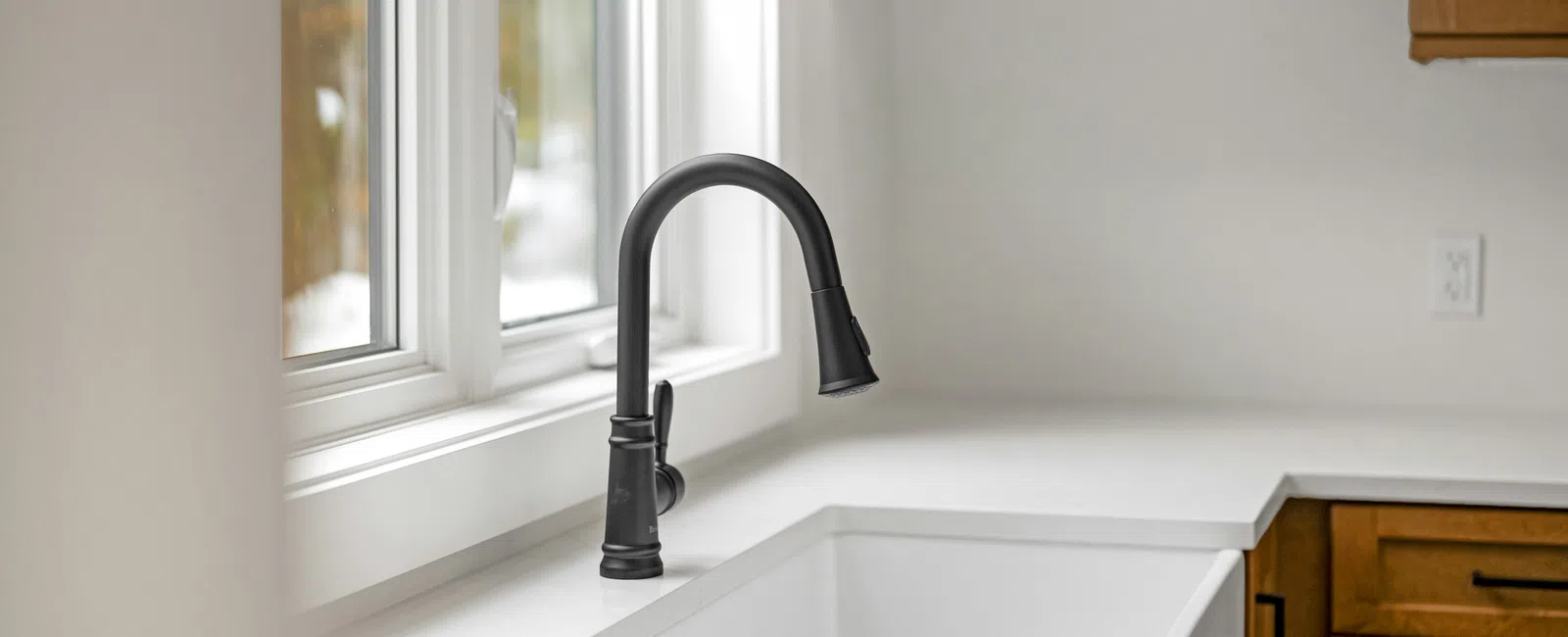 Modern kitchen sink with matte black faucet, white countertop, and wooden cabinets under natural light.