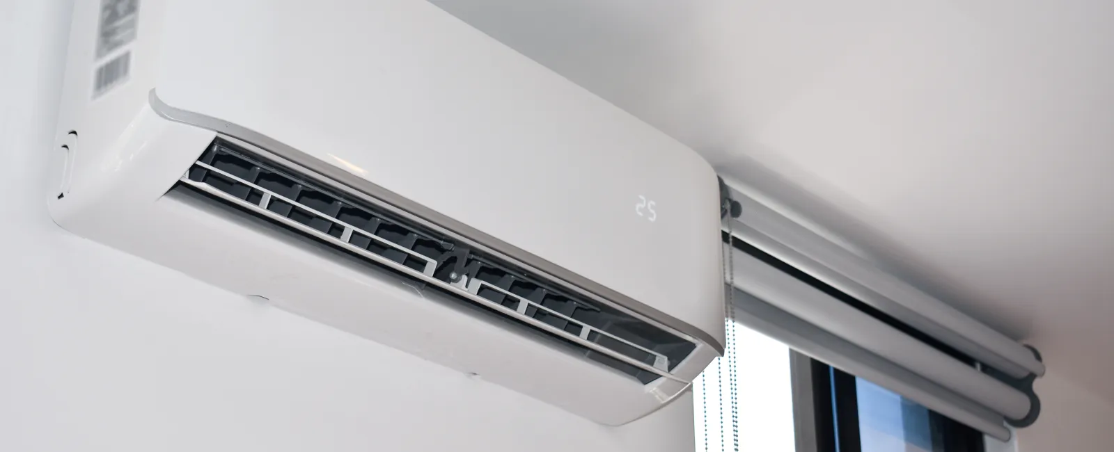 White wall-mounted air conditioner unit showing temperature set to 25 degrees near window with blinds.