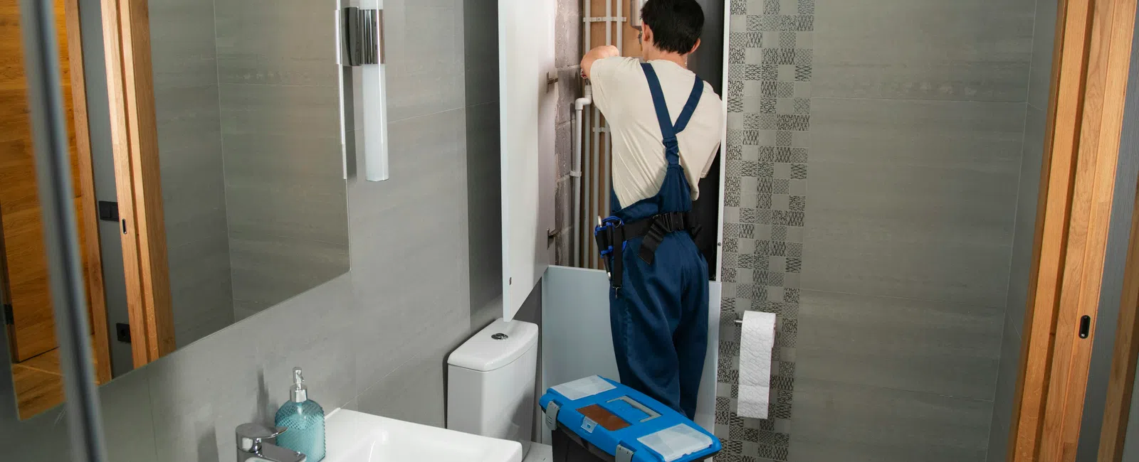 Plumber in blue overalls repairing pipes in a modern bathroom with gray tiles and white fixtures.