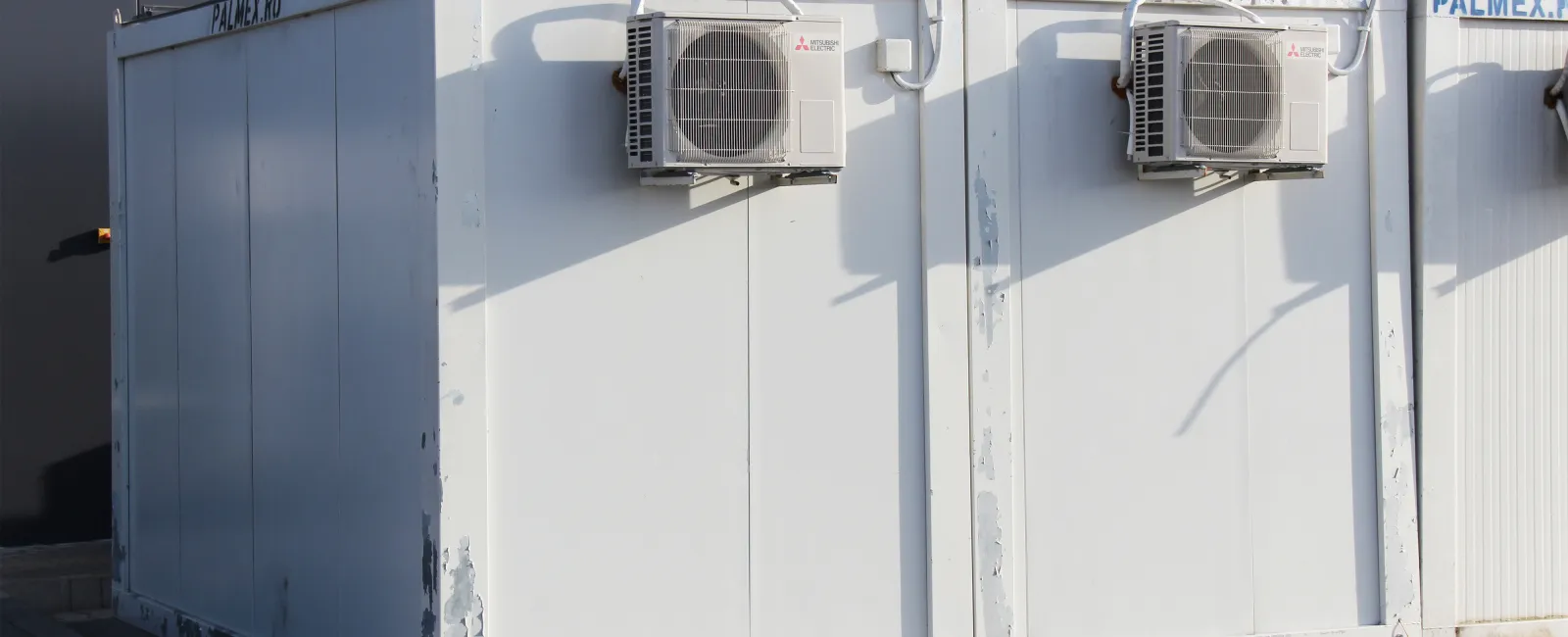 Two white metal containers with mounted air conditioning units on a paved outdoor surface in daylight