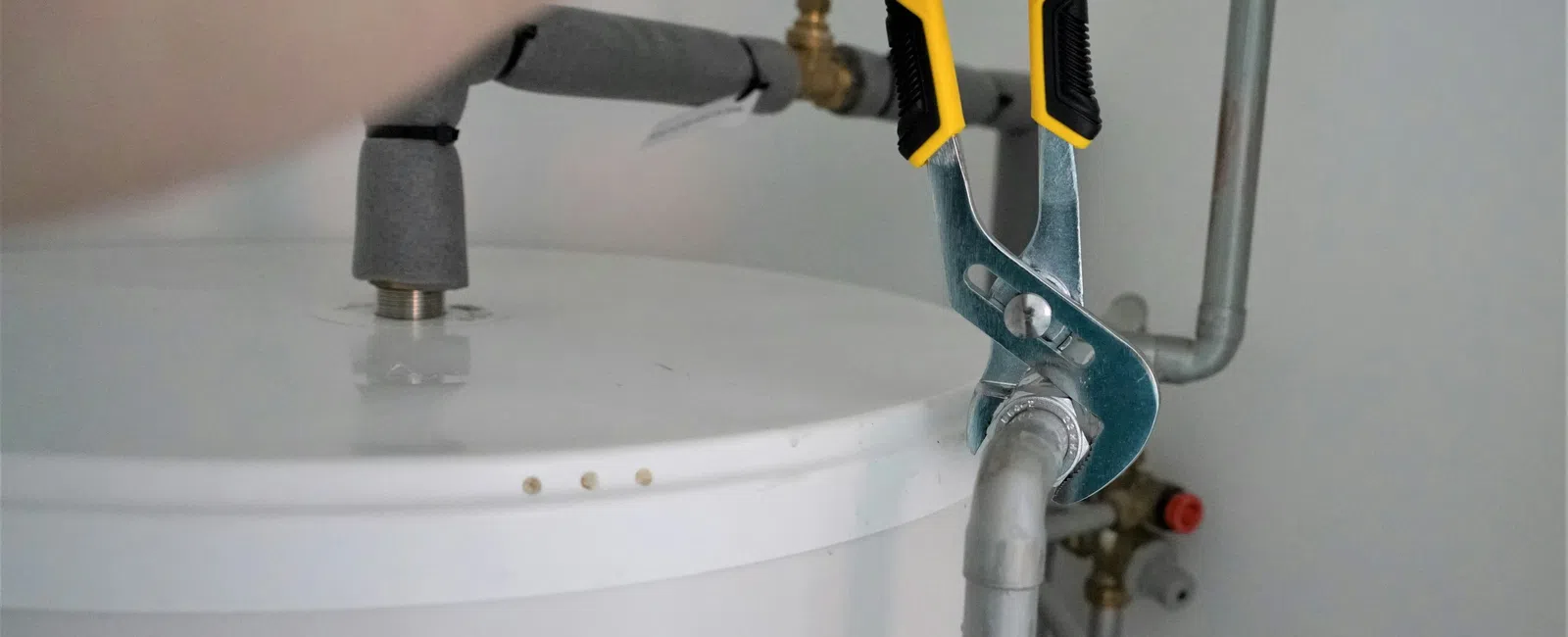 Hand using yellow pliers to tighten pipe connected to a white water heater with visible labels and valves.