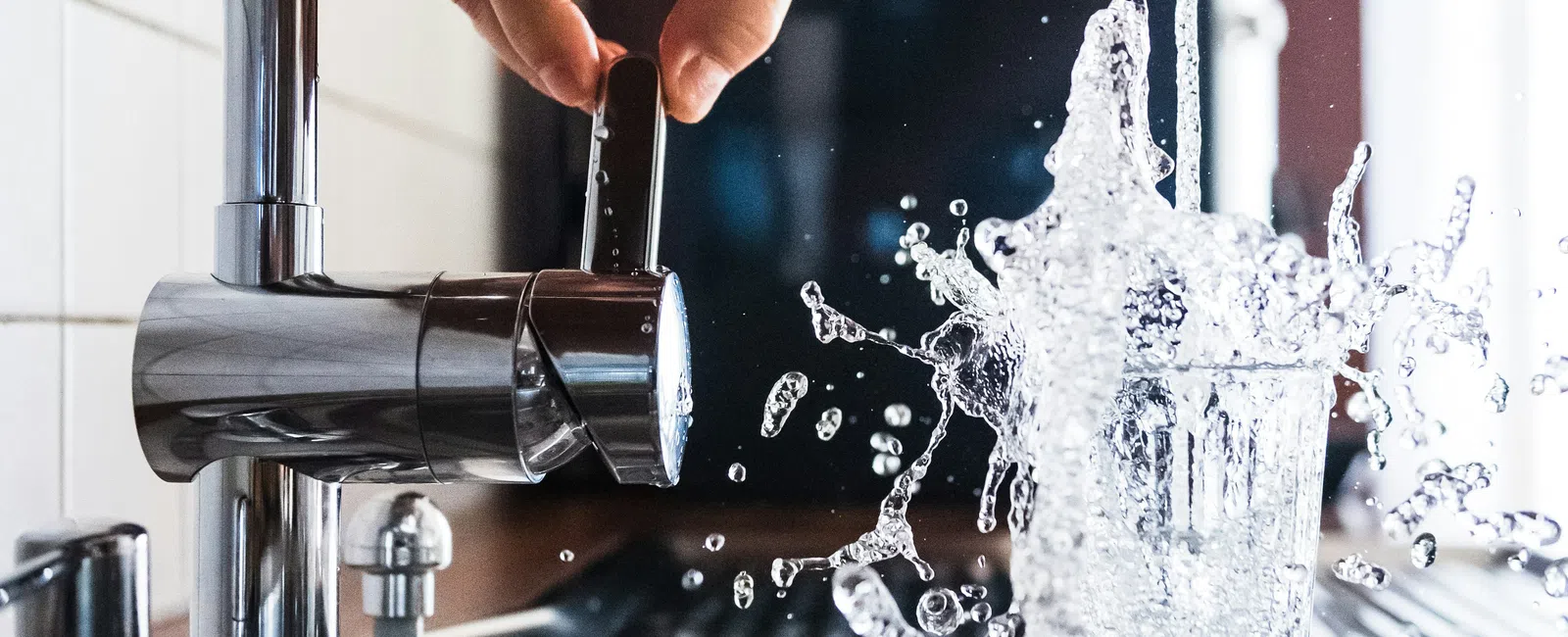 Hand turning faucet on as water splashes out of a glass in a kitchen sink, motion captured mid-splash.