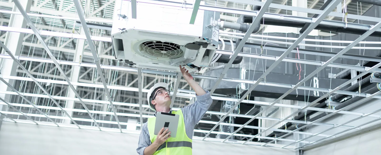 Technician inspecting ceiling HVAC unit, holding tablet, wearing safety vest and helmet in industrial building.