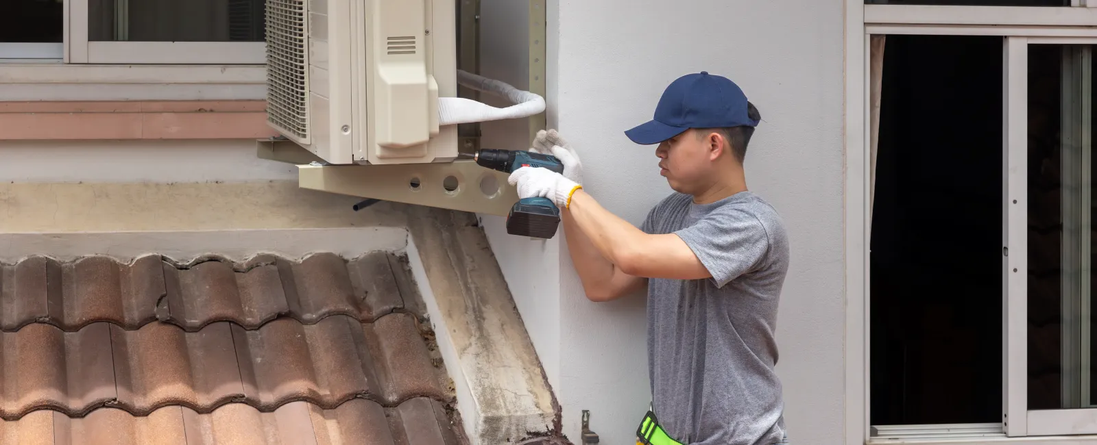 Technician wearing gloves and cap installs an air conditioning unit on a house exterior wall above a tiled roof.