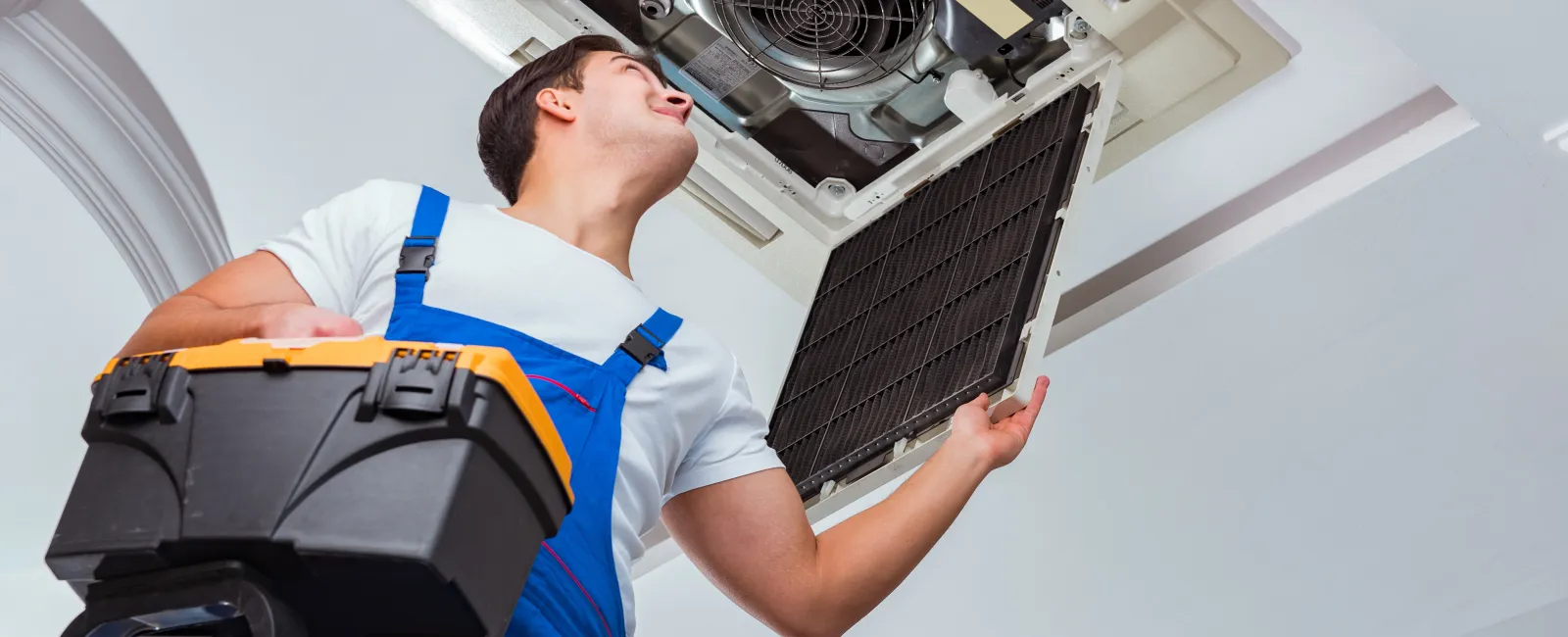 Technician in blue overalls inspecting and repairing ceiling air conditioning unit while holding a black toolbox