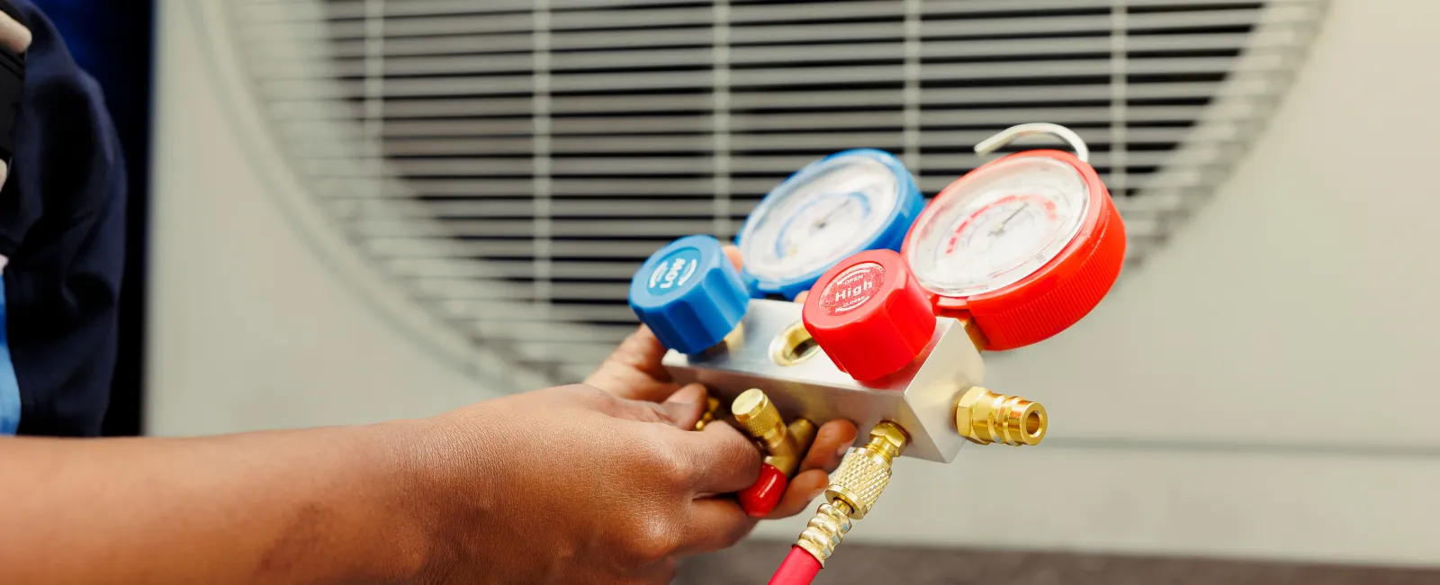 Technician's hand adjusting HVAC manifold gauge with blue and red hoses near an air conditioning unit outdoors.