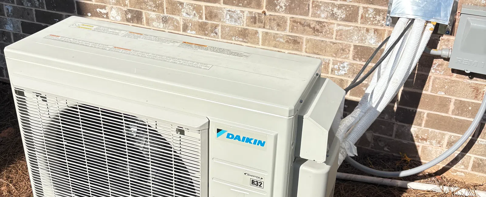 Daikin outdoor air conditioning unit installed on a concrete slab against a brick wall with connected insulated pipes.