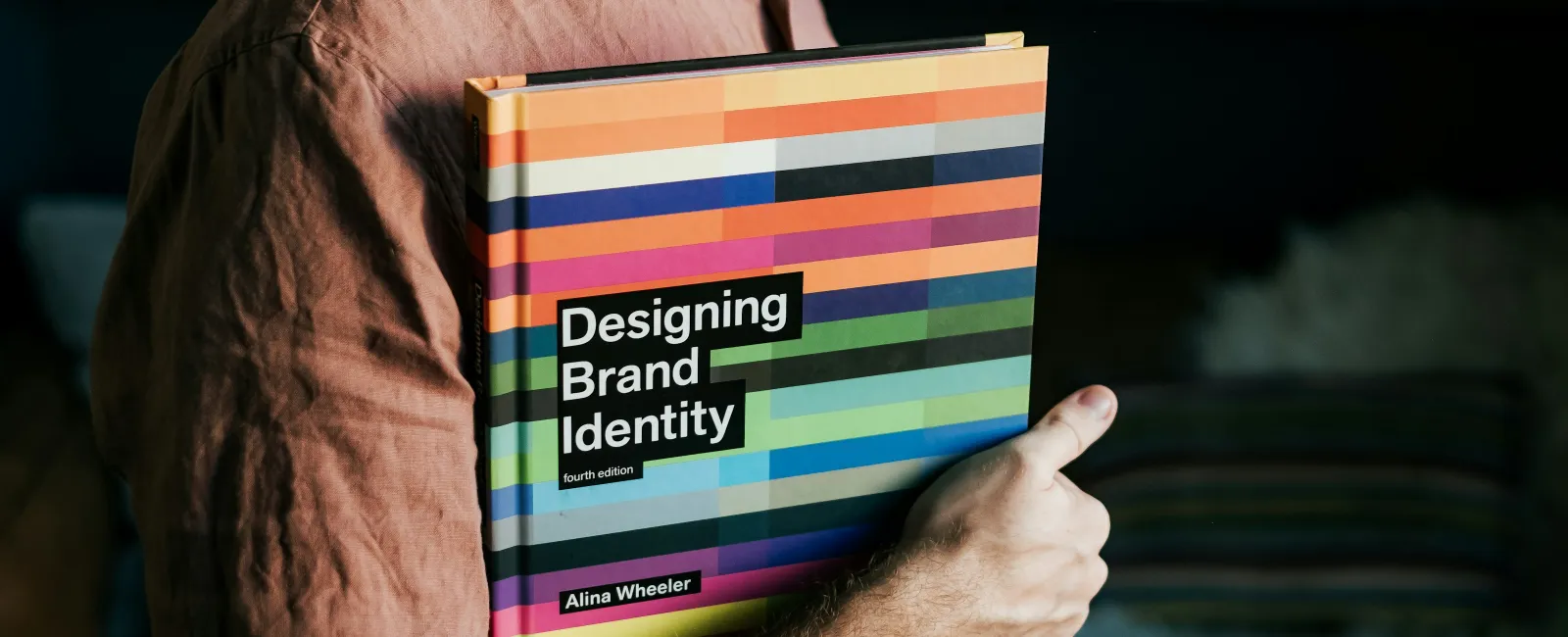 Person holding a colorful book titled Designing Brand Identity by Alina Wheeler in a cozy room.