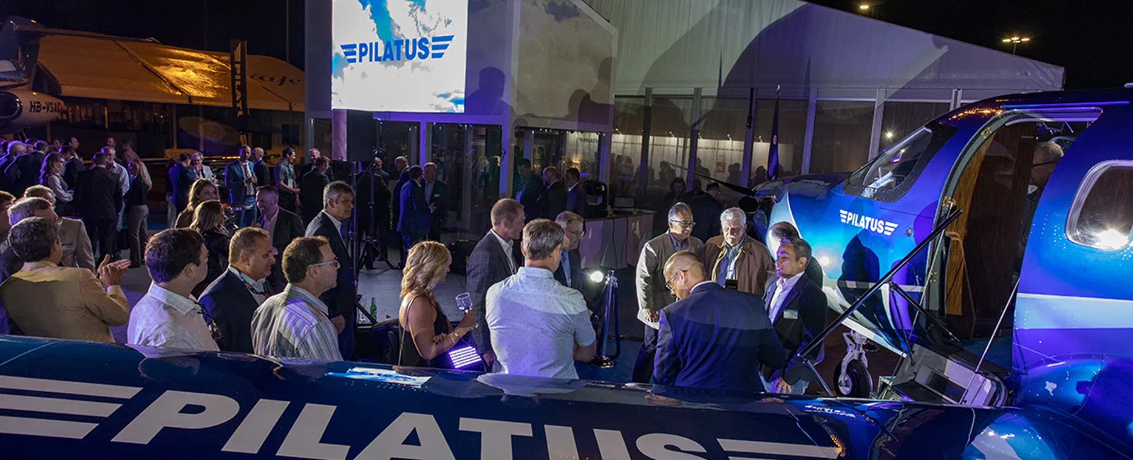 Crowd gathered around Pilatus aircraft at a nighttime event with illuminated signage and displays.