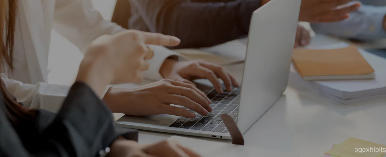 Close-up of hands typing on laptop keyboard with another person pointing at screen during meeting.