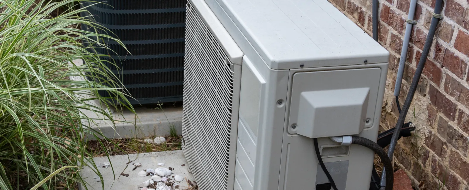 Two outdoor HVAC units on a concrete pad beside a brick house with surrounding green plants and electrical wiring.