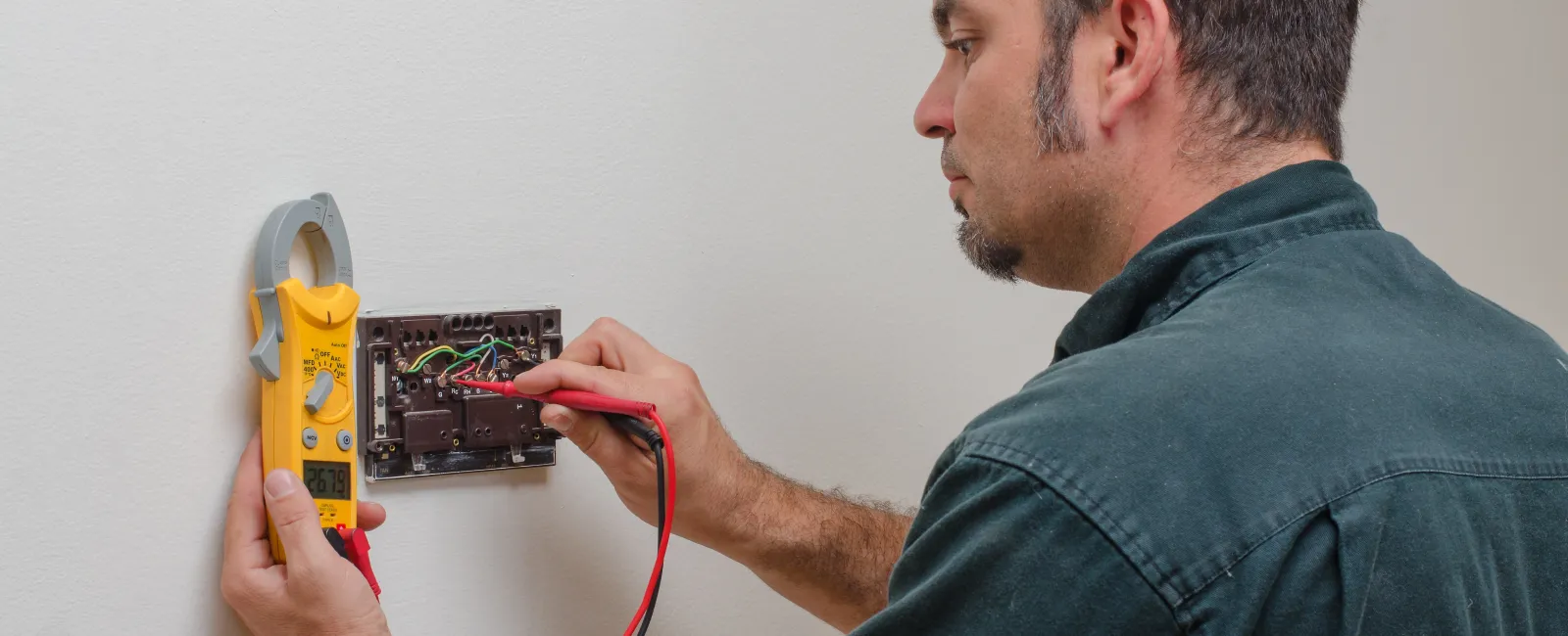 Electrician using a clamp meter to test wiring inside a wall-mounted electrical panel, ensuring safety and functionality