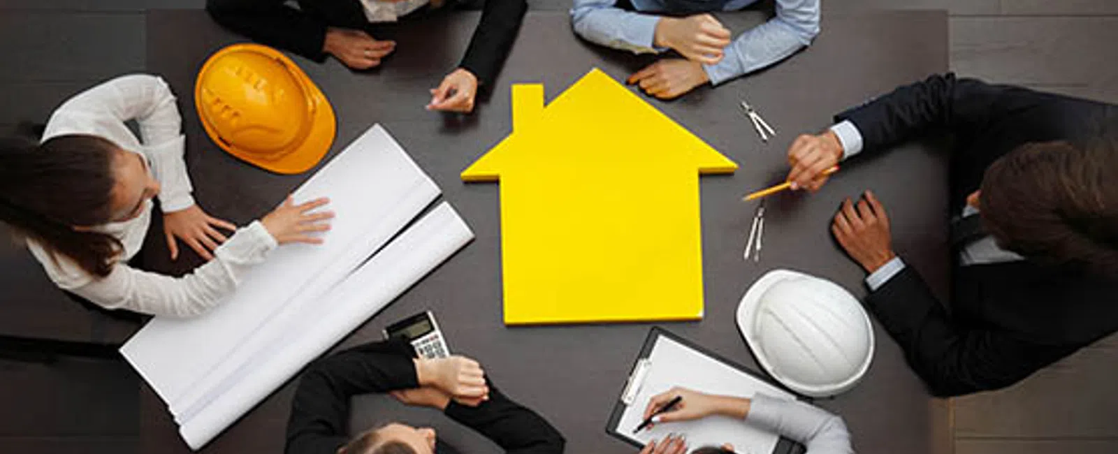 Five professionals discuss construction plans around a table with a yellow house cutout and hard hats.