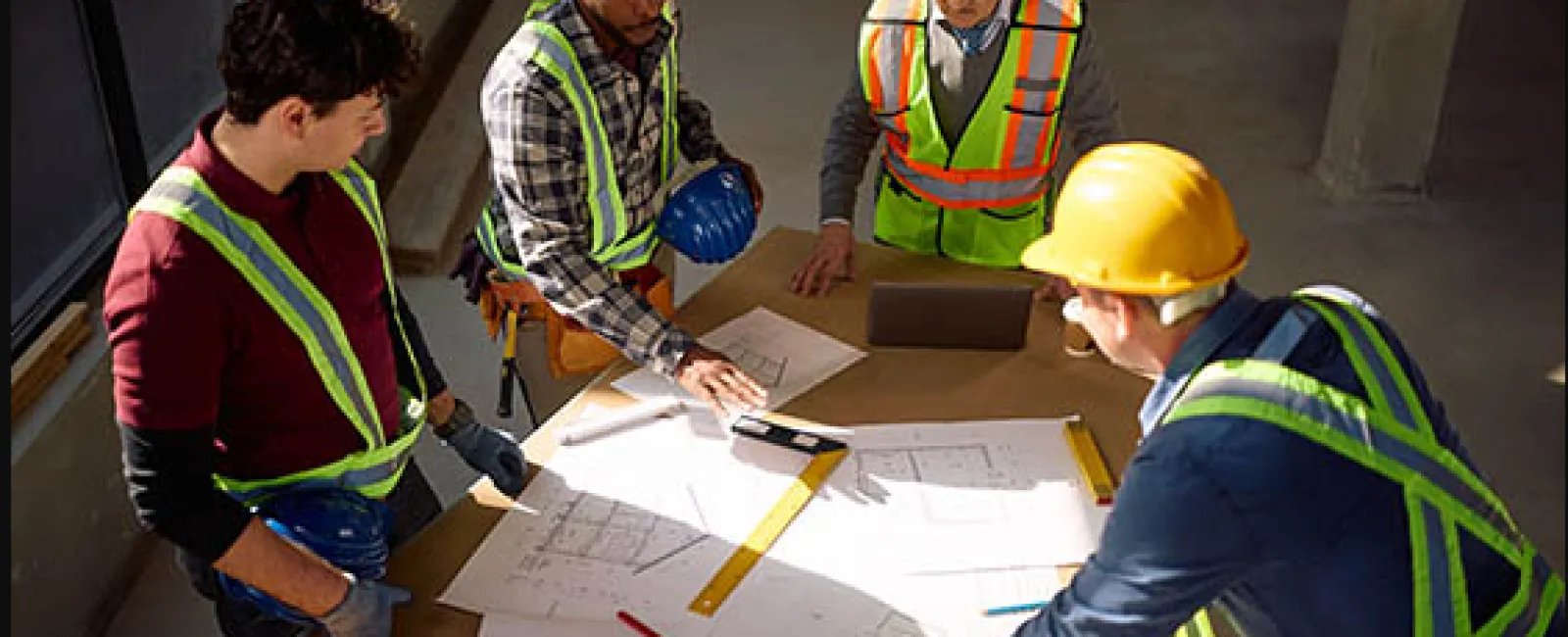 Four construction workers in safety gear reviewing blueprints and plans on a table at a building site.