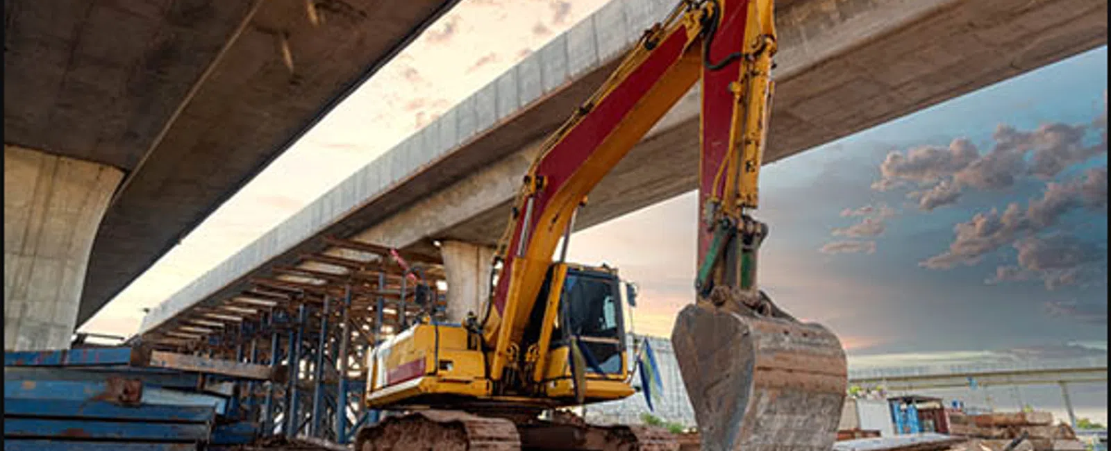 Yellow excavator with red arm working under a concrete bridge at a construction site during sunset.