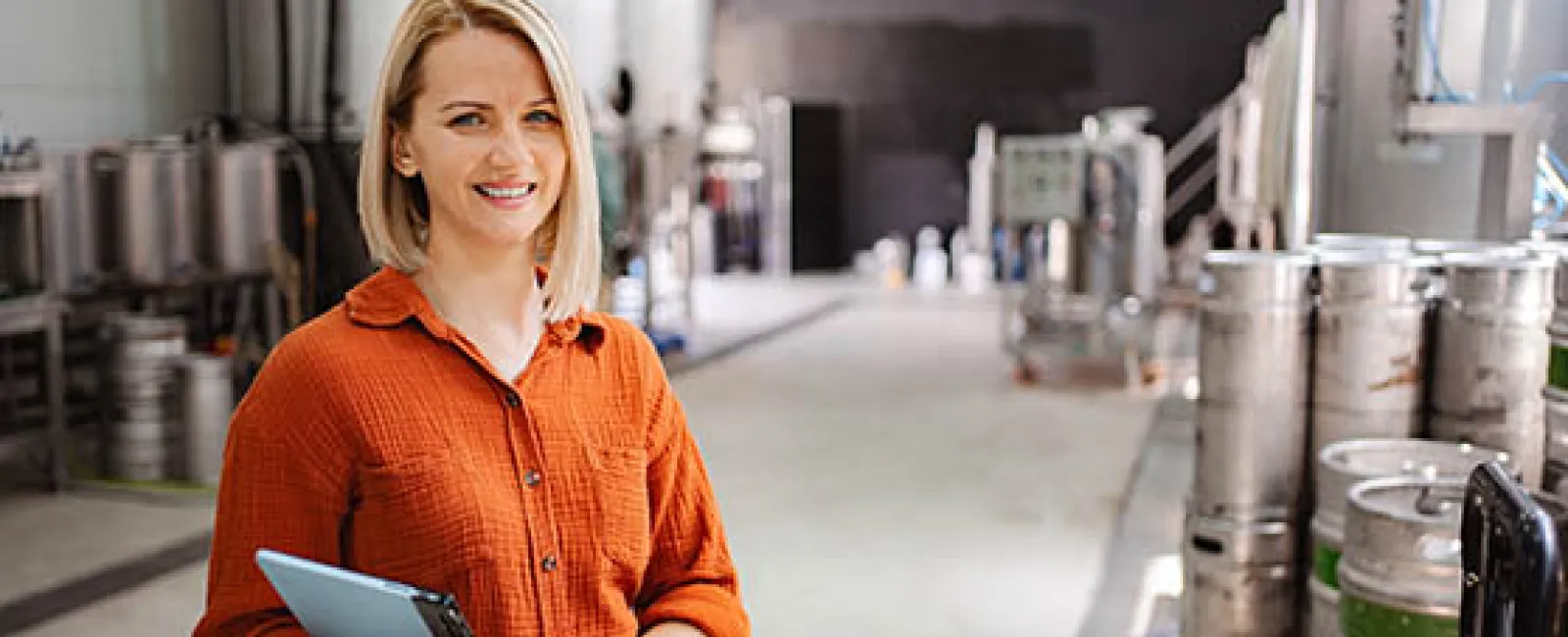 Smiling woman in orange shirt holding tablet stands in brewery surrounded by metal kegs and equipment.