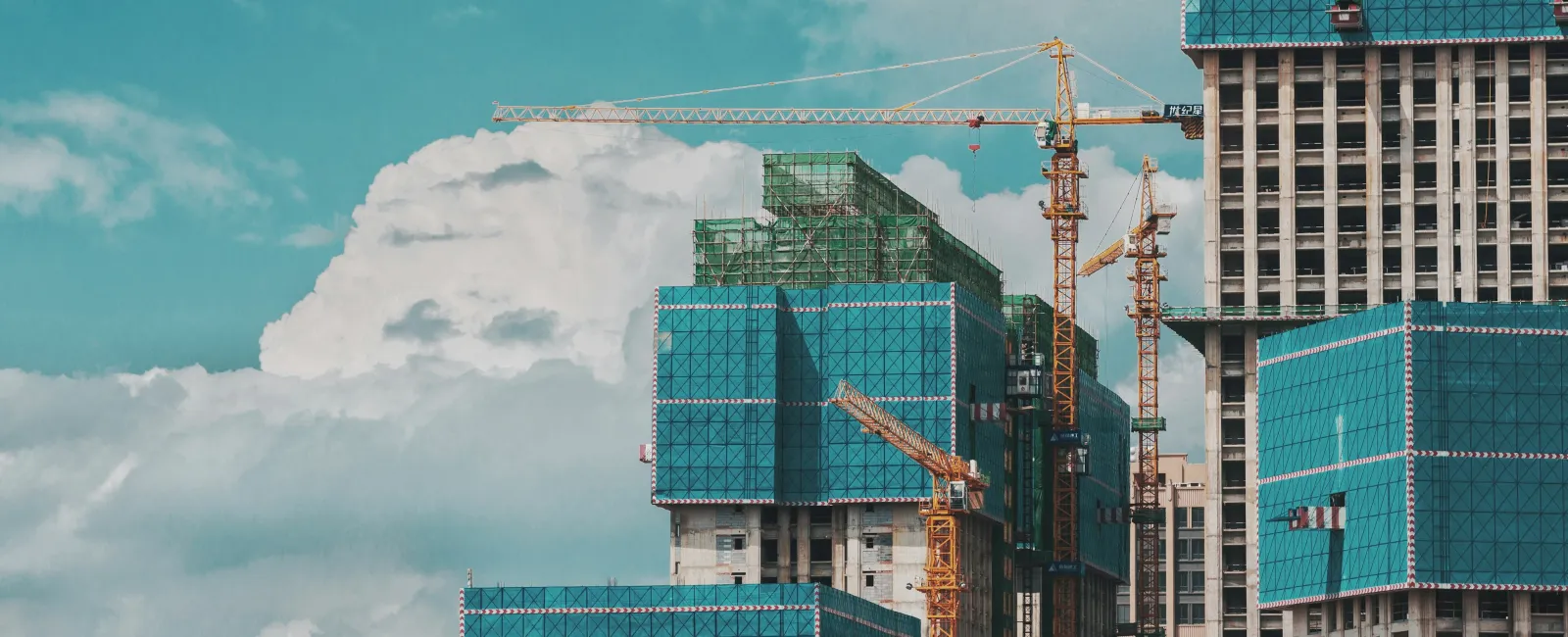 Construction cranes and modern buildings partially covered in blue safety netting against a cloudy sky.