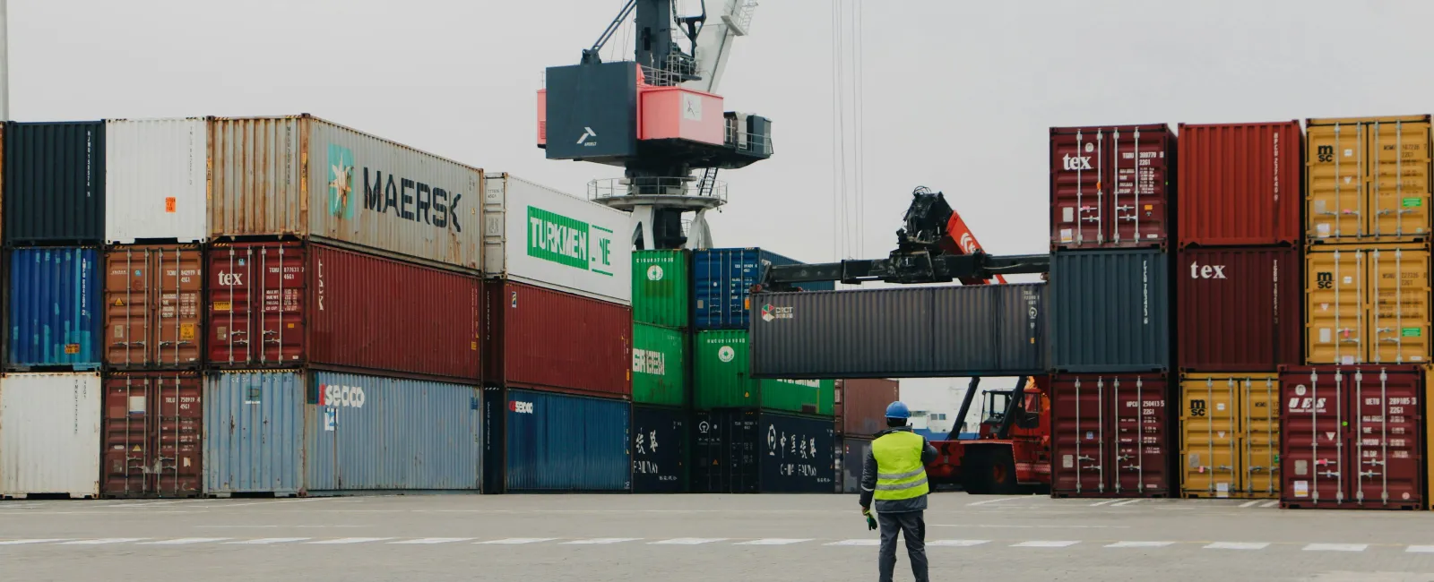 Worker in safety vest directing the loading of shipping containers at a busy industrial port with cranes.