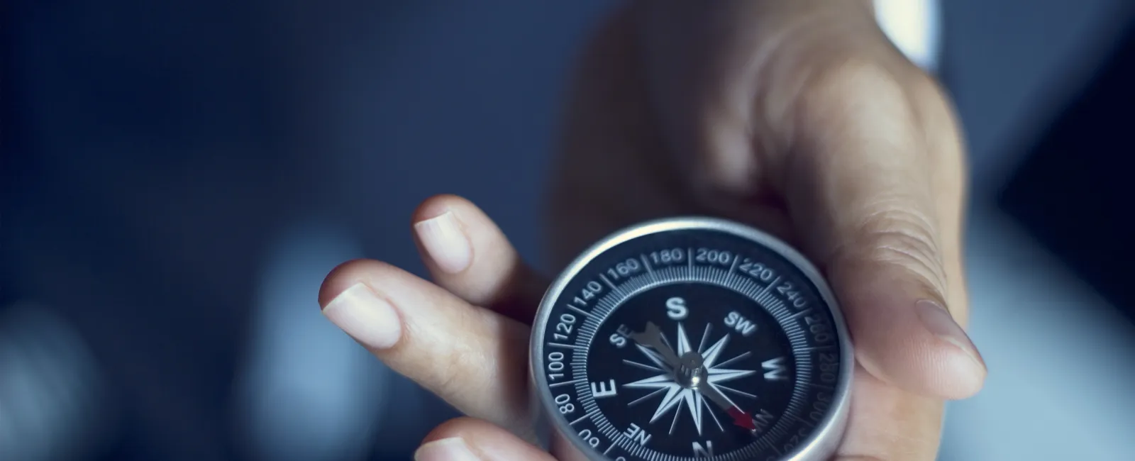 Hand holding a silver compass with clear directional markings against a blurred dark background.