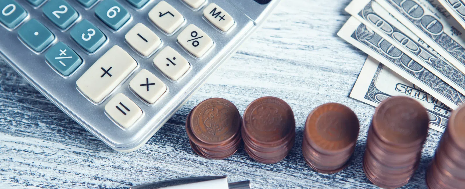 Calculator, stacks of coins, U.S. dollar bills, and a pen on a gray wooden surface representing finance and budgeting