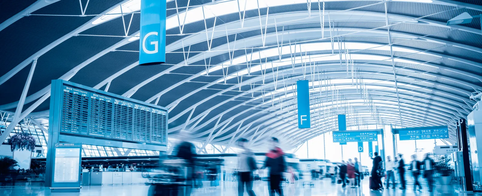 Modern airport terminal with curved ceiling, digital flight boards, and passengers walking with luggage.