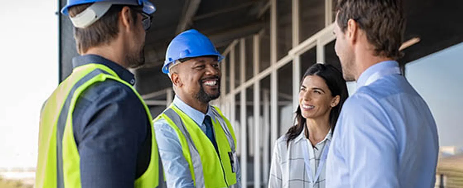 Construction professionals in safety gear shaking hands and smiling outdoors by a modern building under construction.