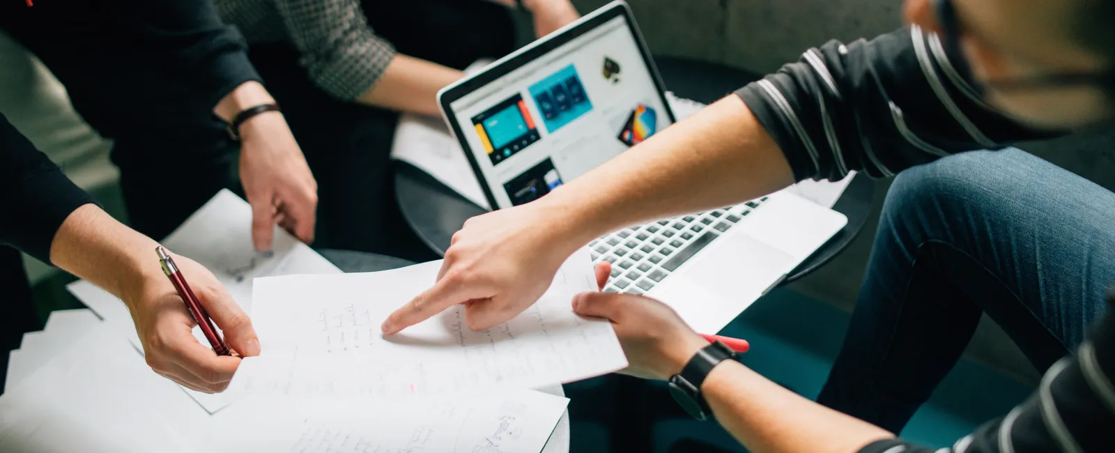 Group of people collaborating on documents with a laptop, discussing ideas and pointing at notes in a casual workspace.