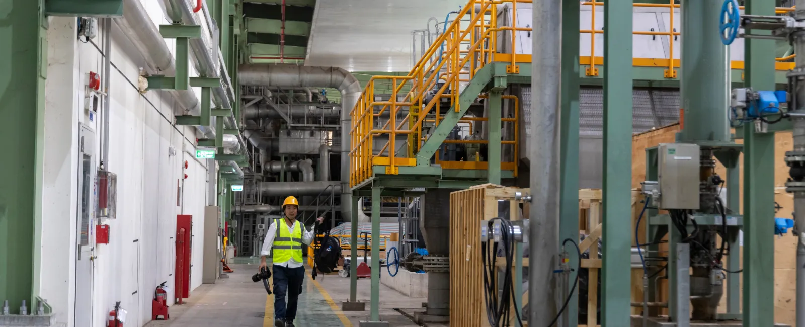 Worker in safety gear walking inside modern factory with machinery and yellow overhead crane