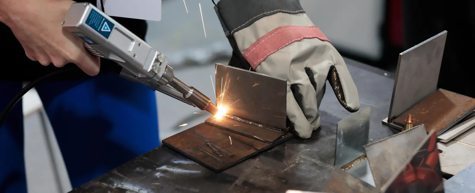 Person using a welding tool to join metal pieces on a workbench with protective gloves.