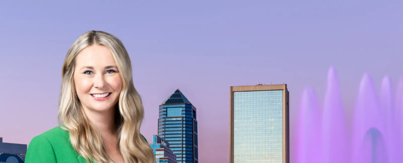 Smiling woman in green blazer stands by purple-lit fountain with Jacksonville skyline at sunset in the background.