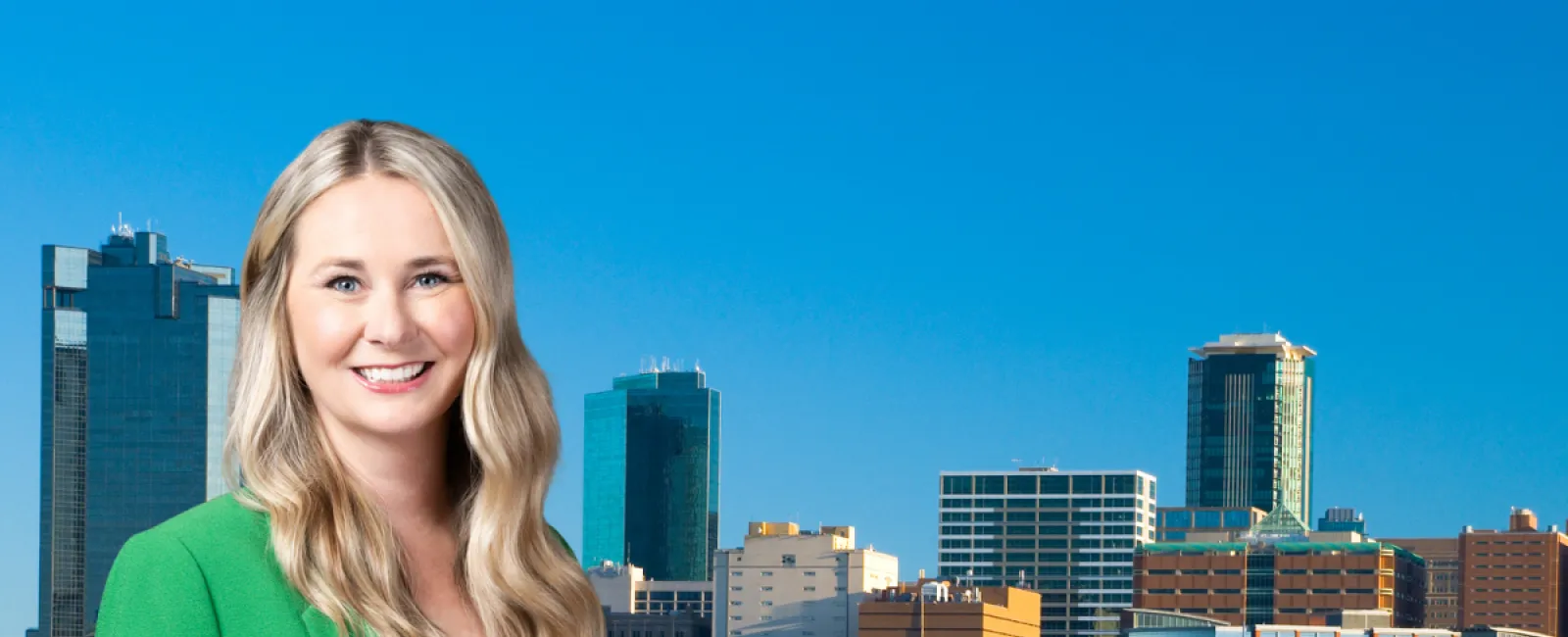 Smiling woman in green blazer with Fort Worth, TX skyline reflected in lake behind her under clear blue sky