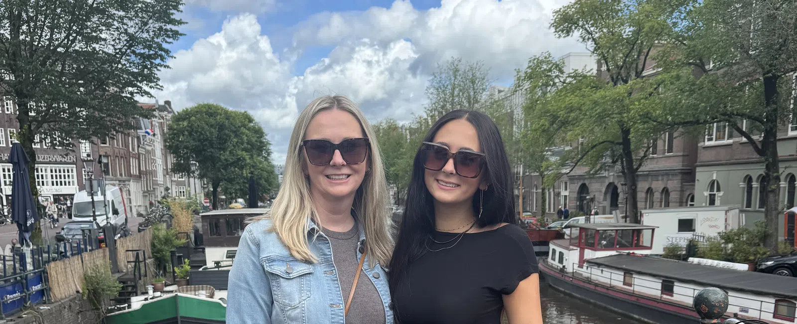 Two women in casual outfits and sunglasses standing by a canal with boats and trees in the background on a sunny day.
