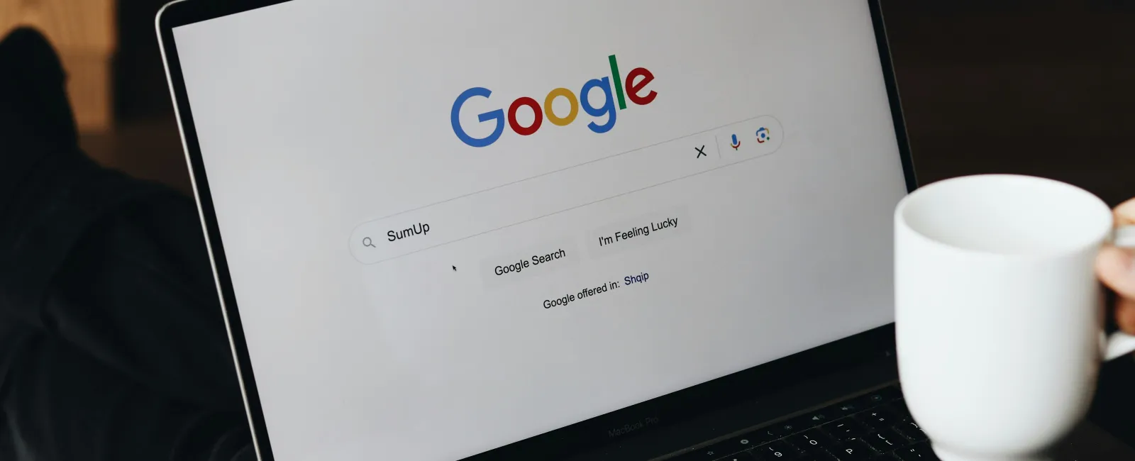 Person using a laptop with Google search page open and holding a white coffee mug.