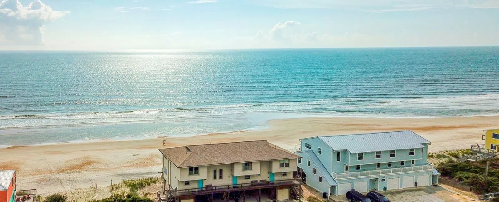 Aerial view of beach houses along a coastal road with a sandy beach and ocean waves under a partly cloudy sky.