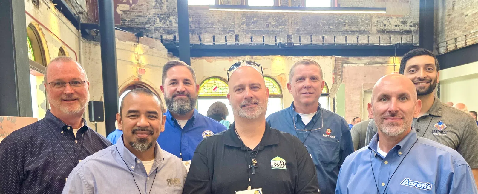 Group of seven men wearing business casual attire and name tags posing inside a rustic industrial building.