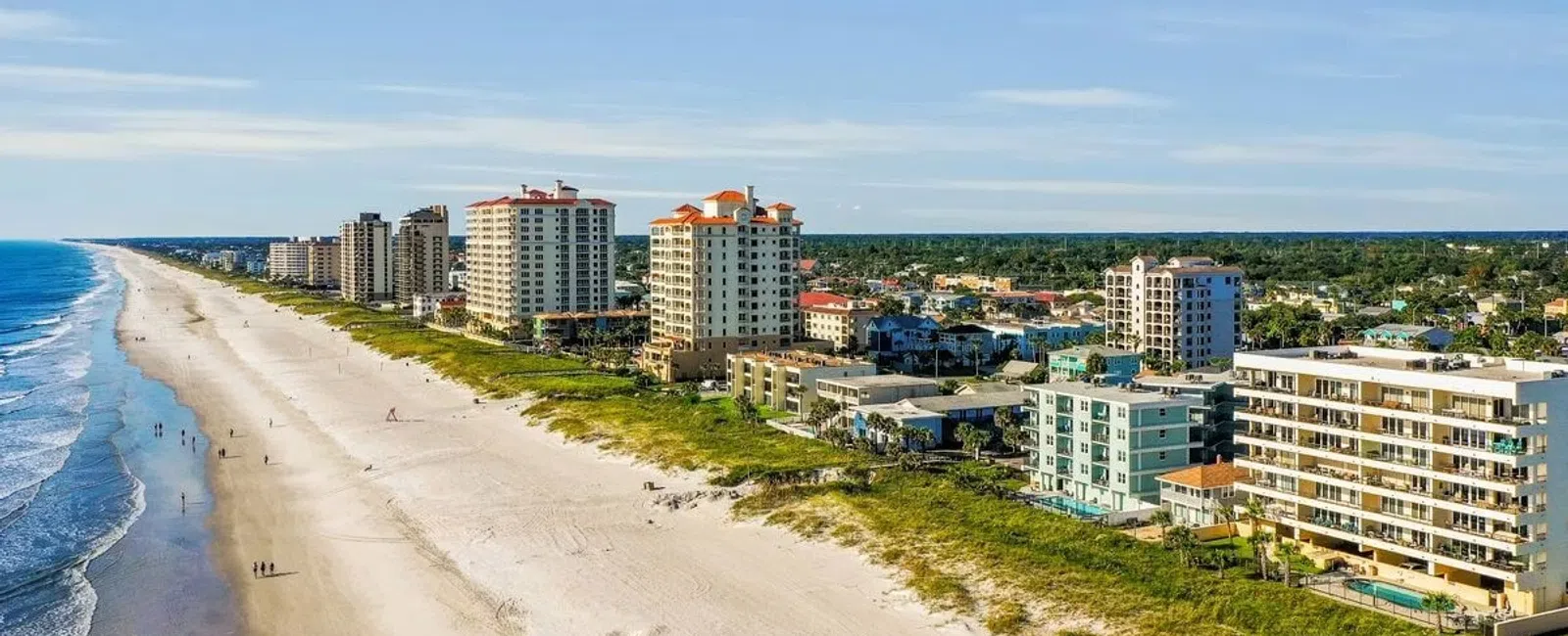Aerial view of a sandy beach with waves and beachfront high-rise buildings under a clear blue sky.