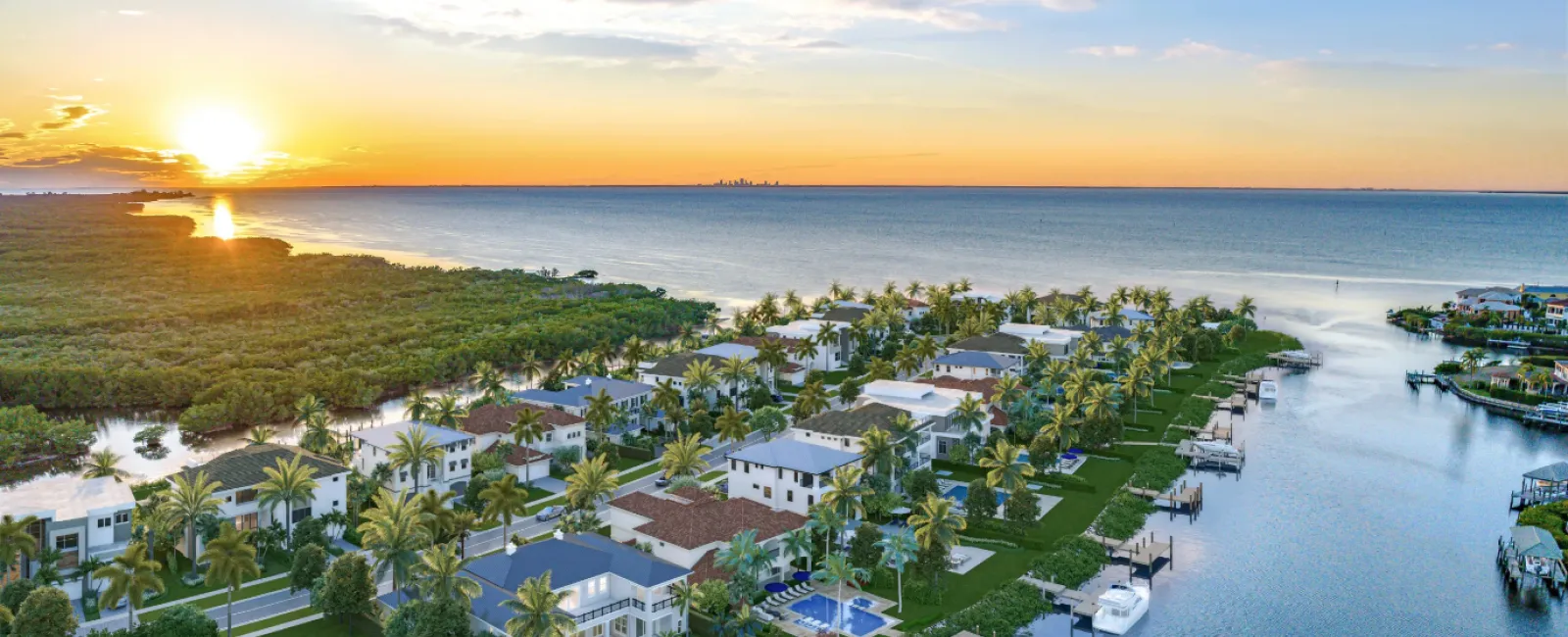 Sunset over modern waterfront homes with palm trees and boats along a canal near the ocean coastline.