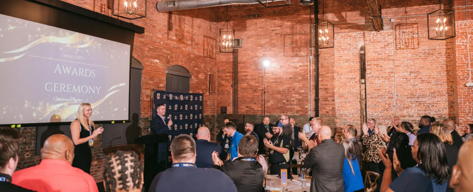 Audience applauding a speaker during an awards ceremony in a rustic brick event space with chandeliers.