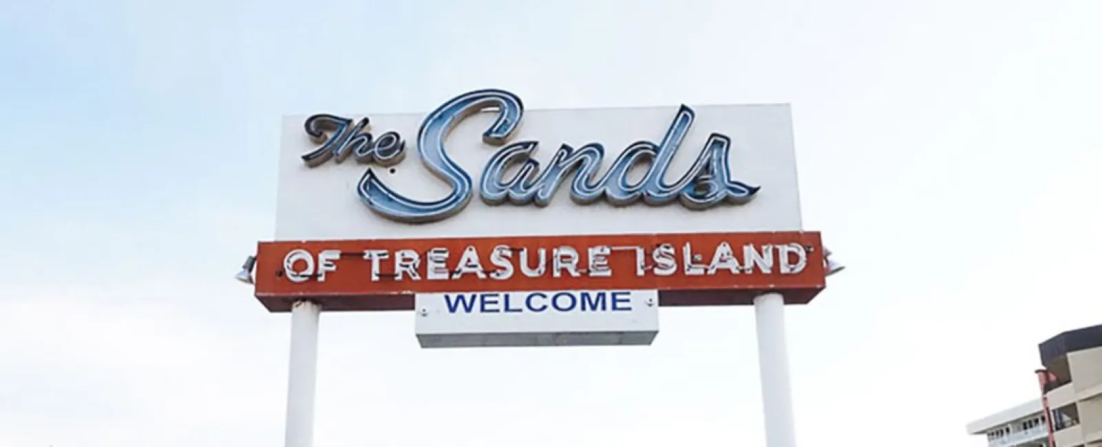 Entrance sign for The Sands of Treasure Island motel with yellow umbrellas and pool chairs under a clear sky