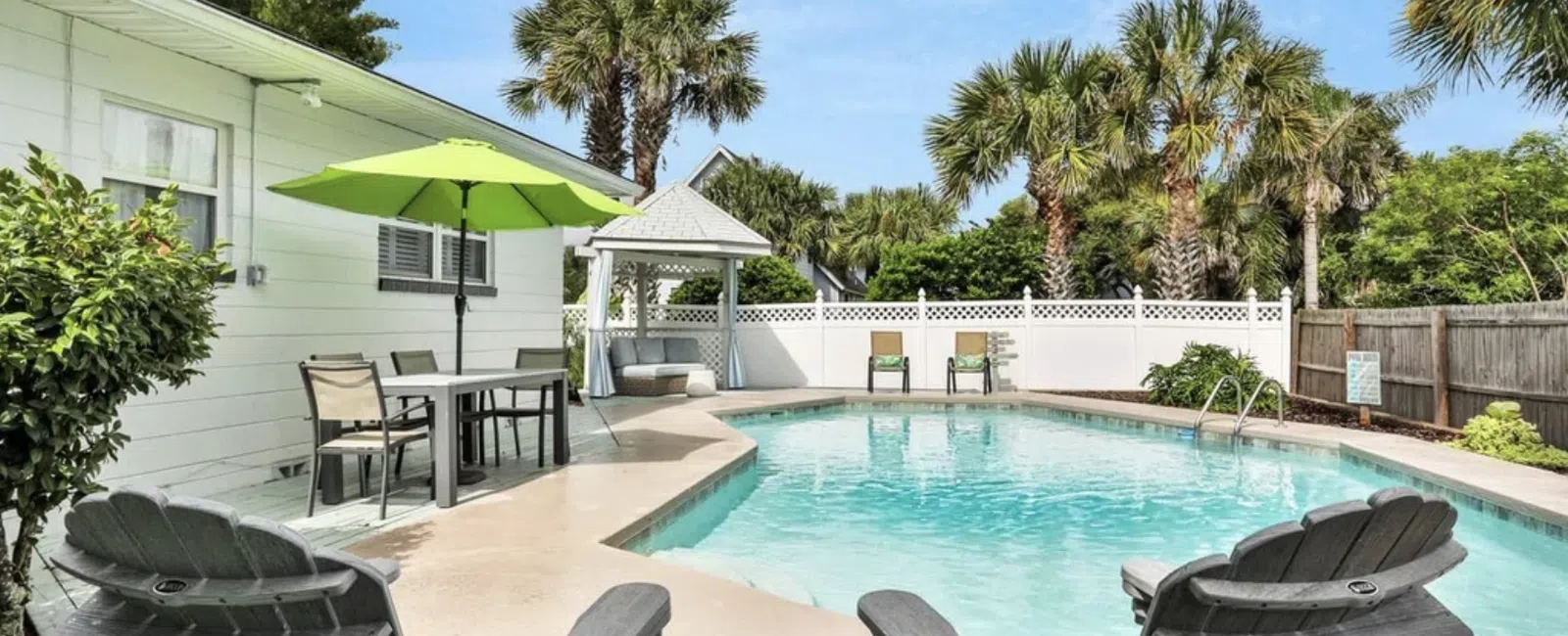 Sunny backyard pool area with palm trees, lounge chairs, green umbrella, and white gazebo under blue sky.