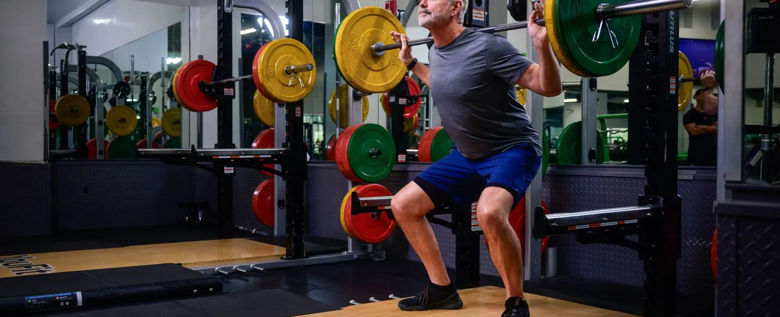 Mature man doing weighted squats with barbell and colorful weights in a modern gym with mirrors.