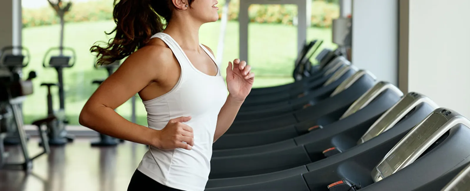 Young woman running on treadmill in a bright gym with multiple exercise machines and large windows.