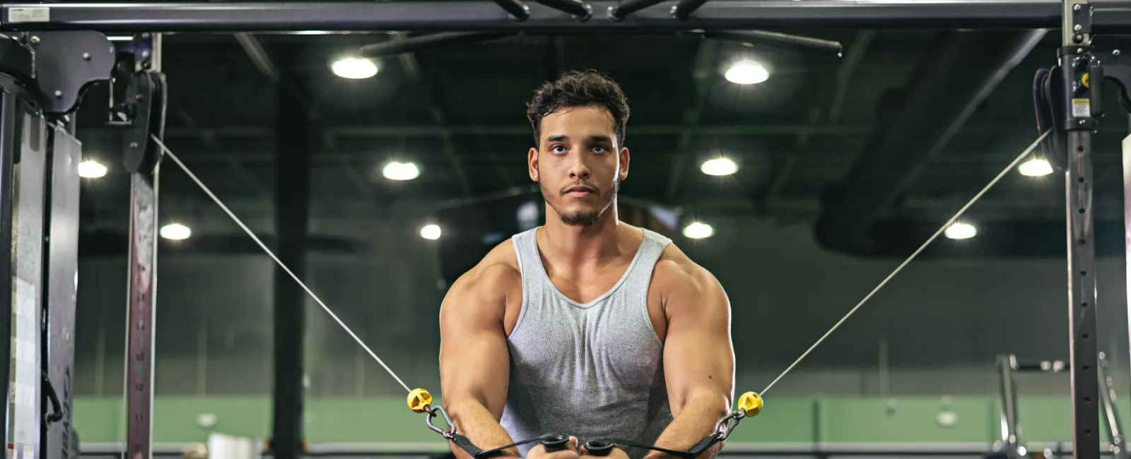 Muscular man in gray tank top and shorts performing cable chest fly exercise in gym
