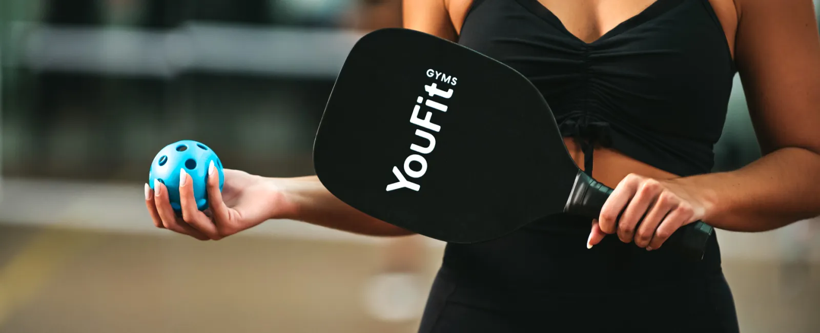 Woman in black sportswear holding a YouFit gym paddle and blue pickleball ball outdoors.