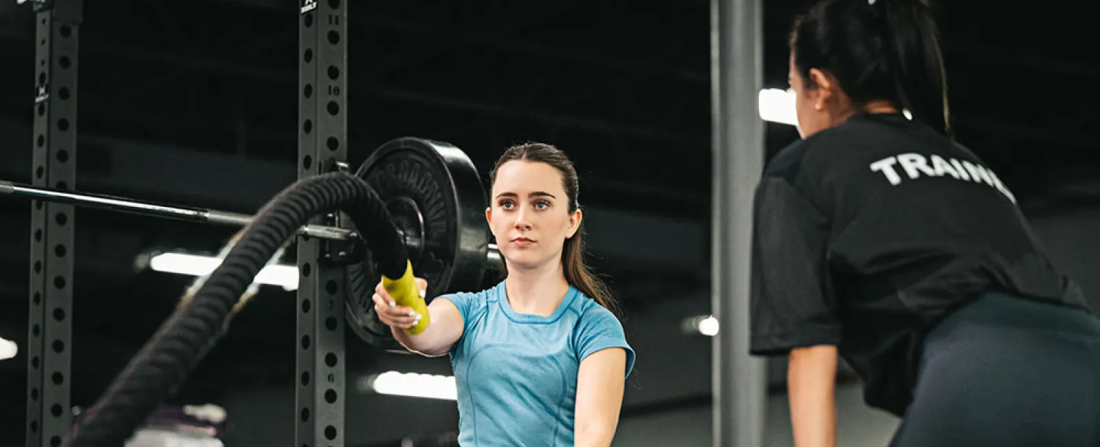 Woman in blue shirt exercises with battle ropes under guidance of female trainer in gym setting