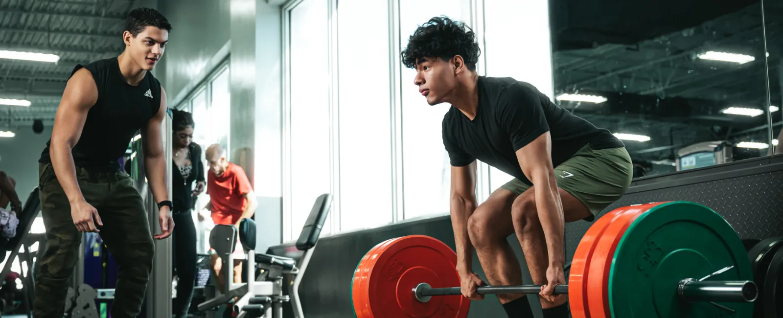 Young man deadlifting heavy barbell at gym while trainer watches and motivates him during workout session.