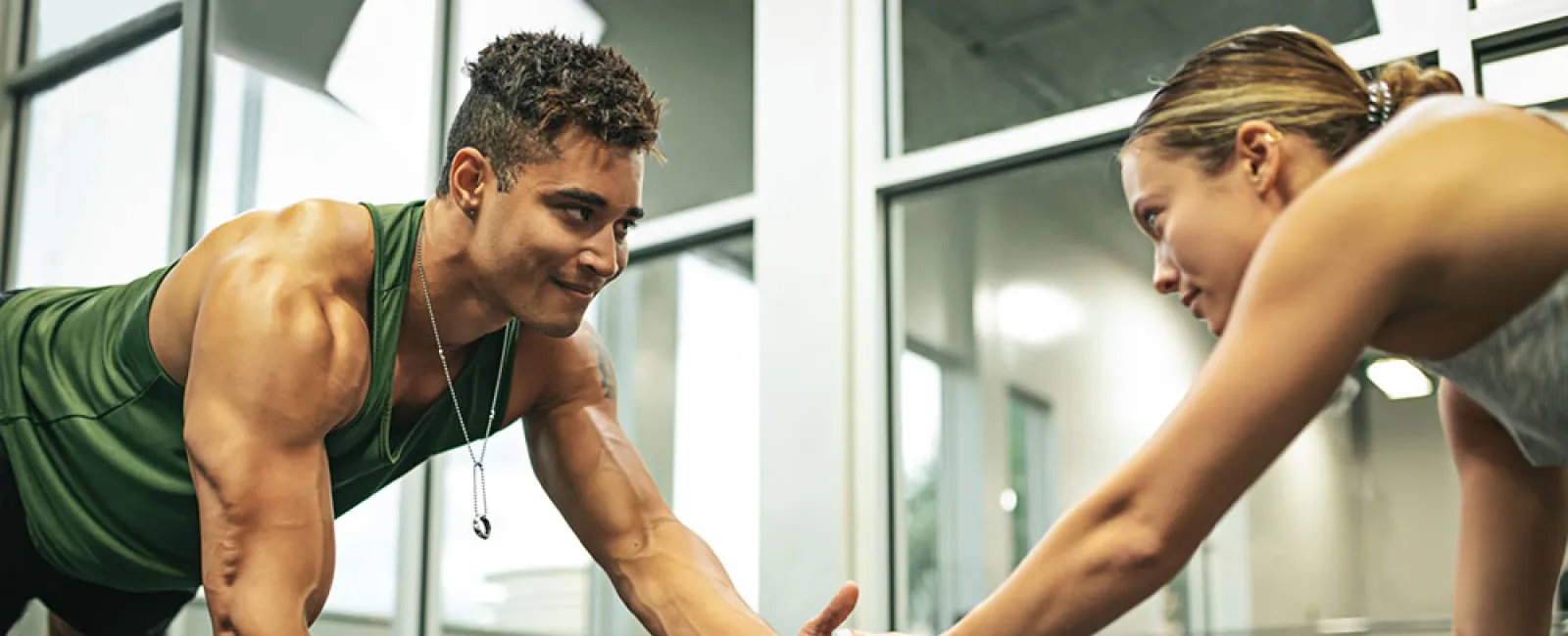 Two fit individuals doing partner plank high-fives in a gym with large windows and modern equipment