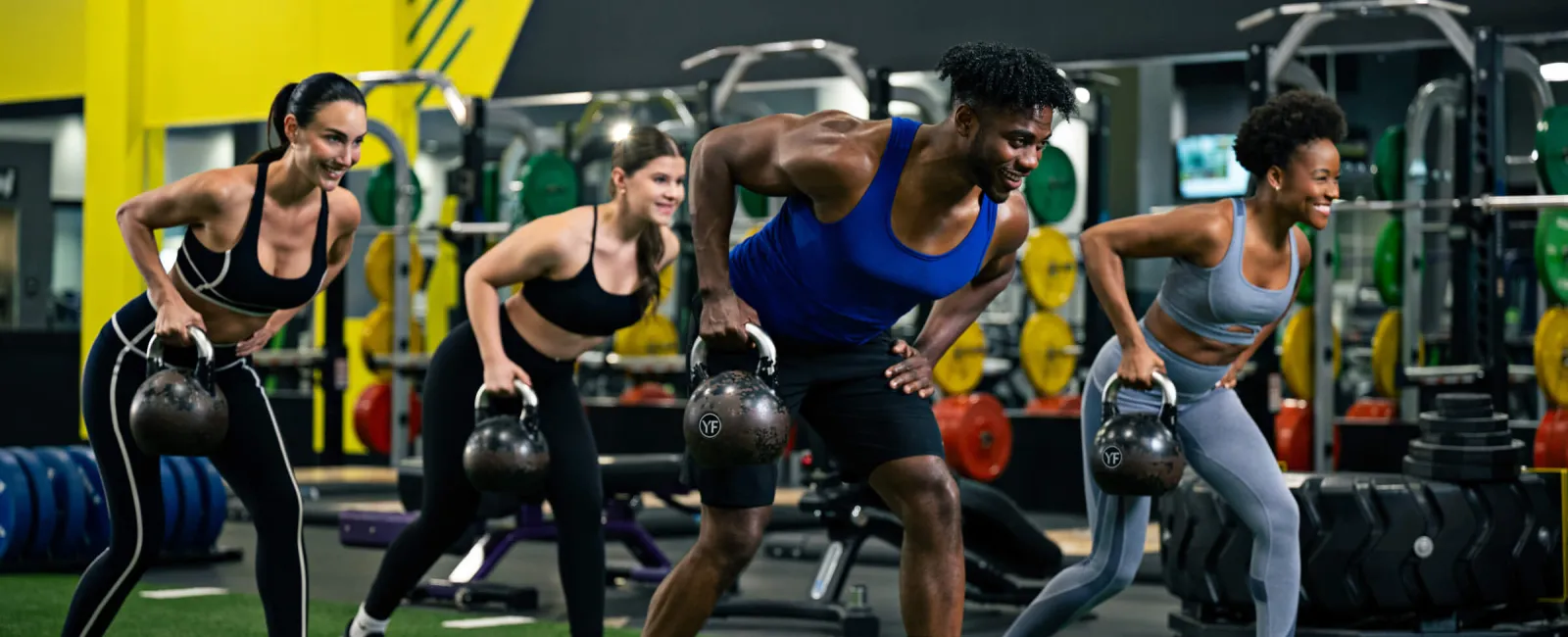 Group of four diverse people exercising with kettlebells in a modern gym with bright yellow walls and equipment.