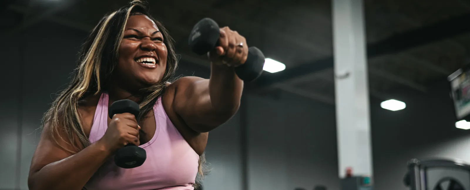 Smiling woman in pink activewear lifting dumbbells and boxing in a gym with equipment in the background